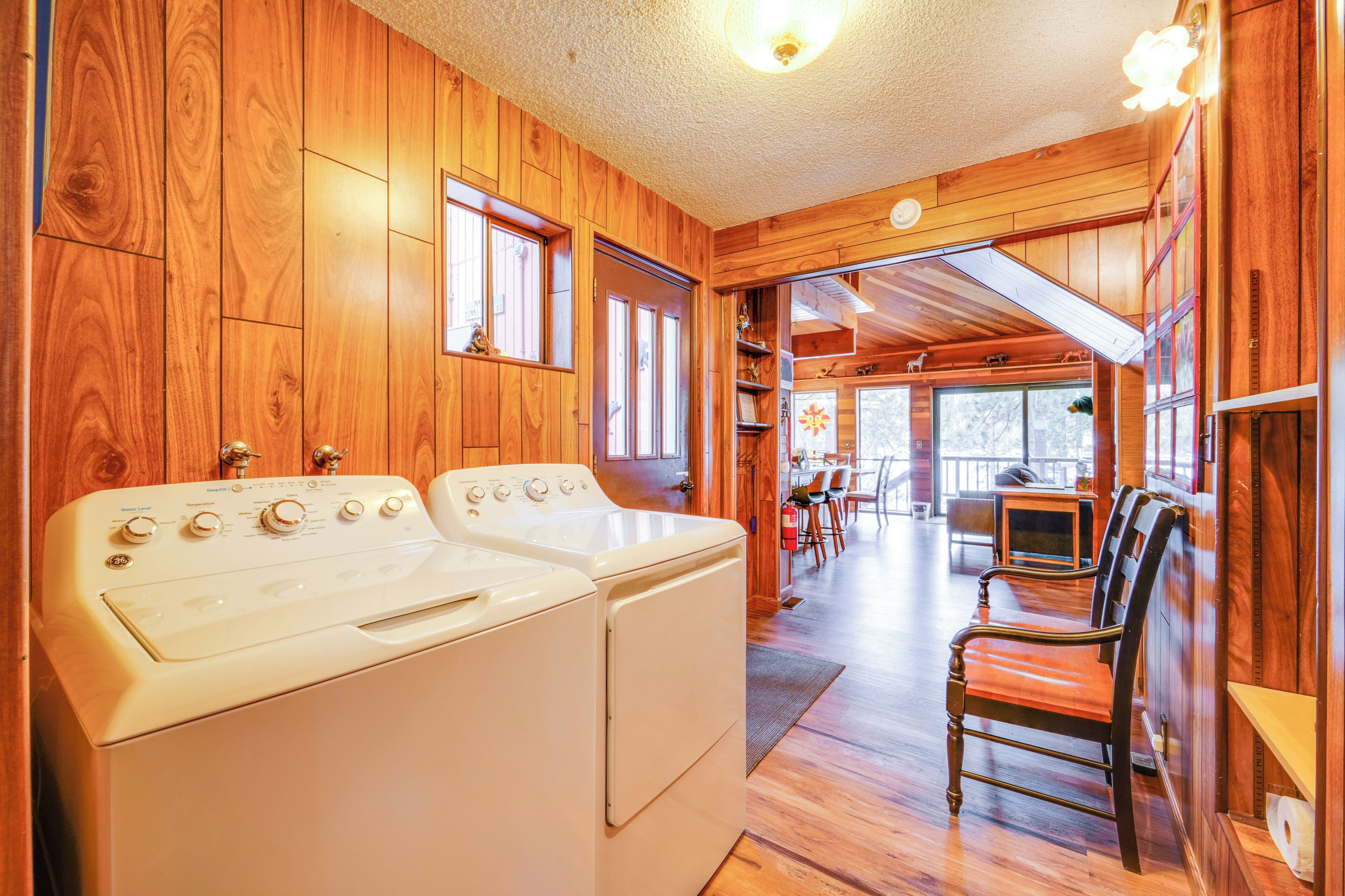A laundry room with two white washing machines, wooden walls, and a view into a dining area with a table and chairs.