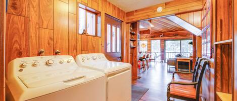 A laundry room with two white washing machines, wooden walls, and a view into a dining area with a table and chairs.