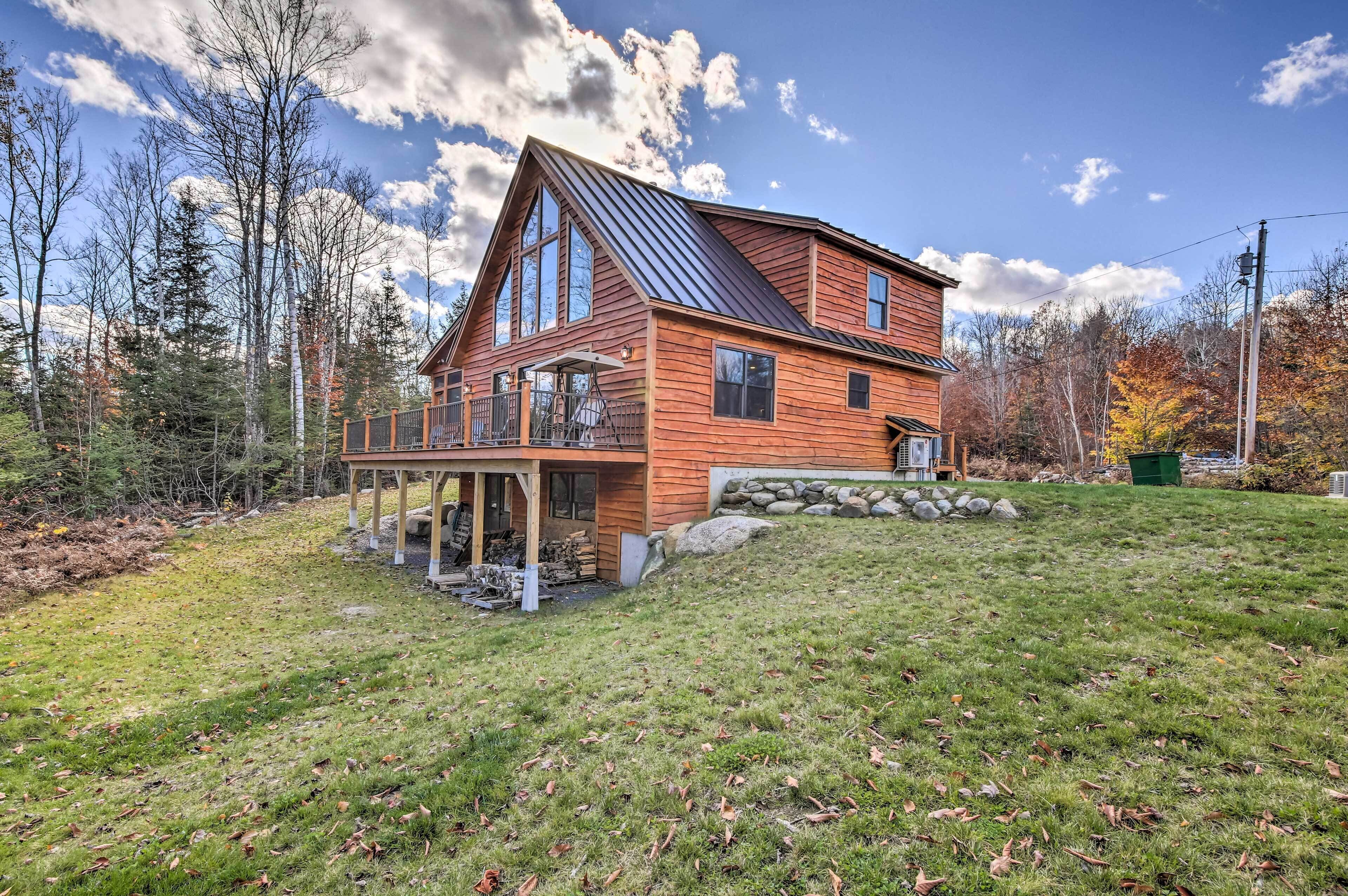 A log cabin with a large deck, surrounded by trees and a grassy yard.