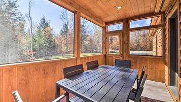 Una terraza de madera con una mesa y sillas, ventanas amplias y vistas a un bosque.