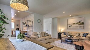 A spacious living area with a wooden dining table, beige sofas, and a coffee table. There is a kitchen area with a refrigerator and a sink in the background.