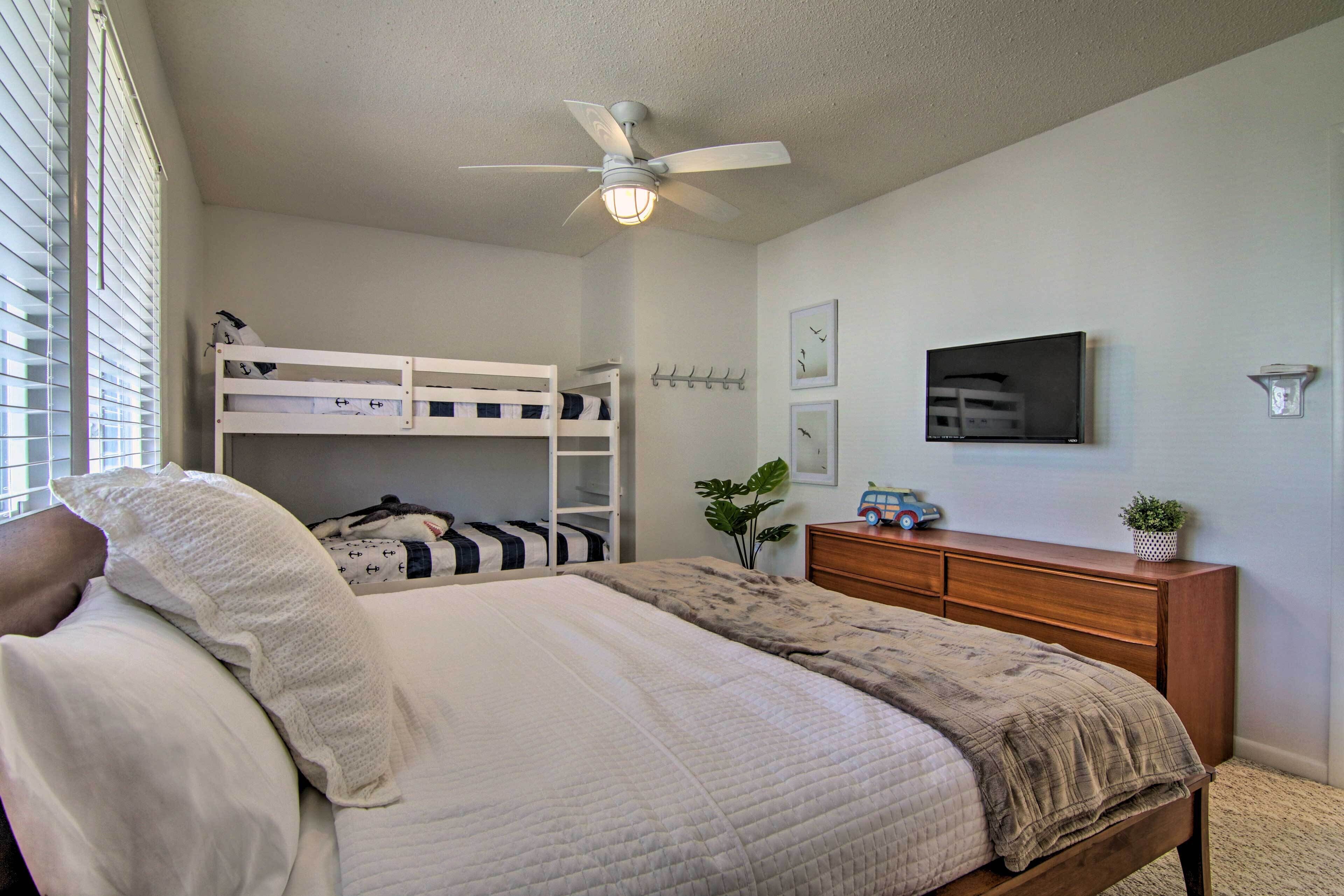 A bedroom with a bunk bed, a single bed, a ceiling fan, a dresser, a television, and wall-mounted hooks.