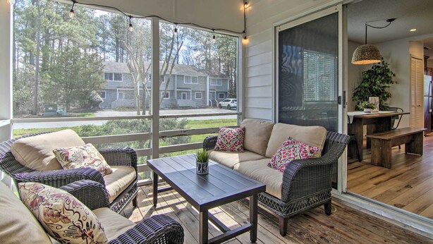 A covered porch with wicker furniture, a table, and a view of houses through large windows.