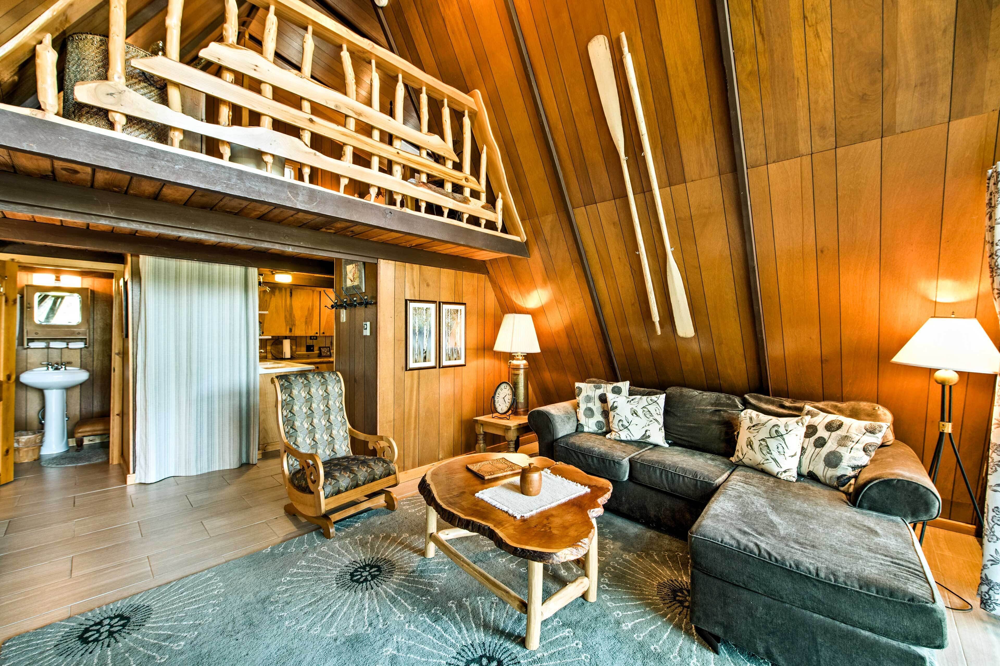 A cozy loft-style room with a wooden ceiling, a sofa, a chair, a small table, and a lamp.