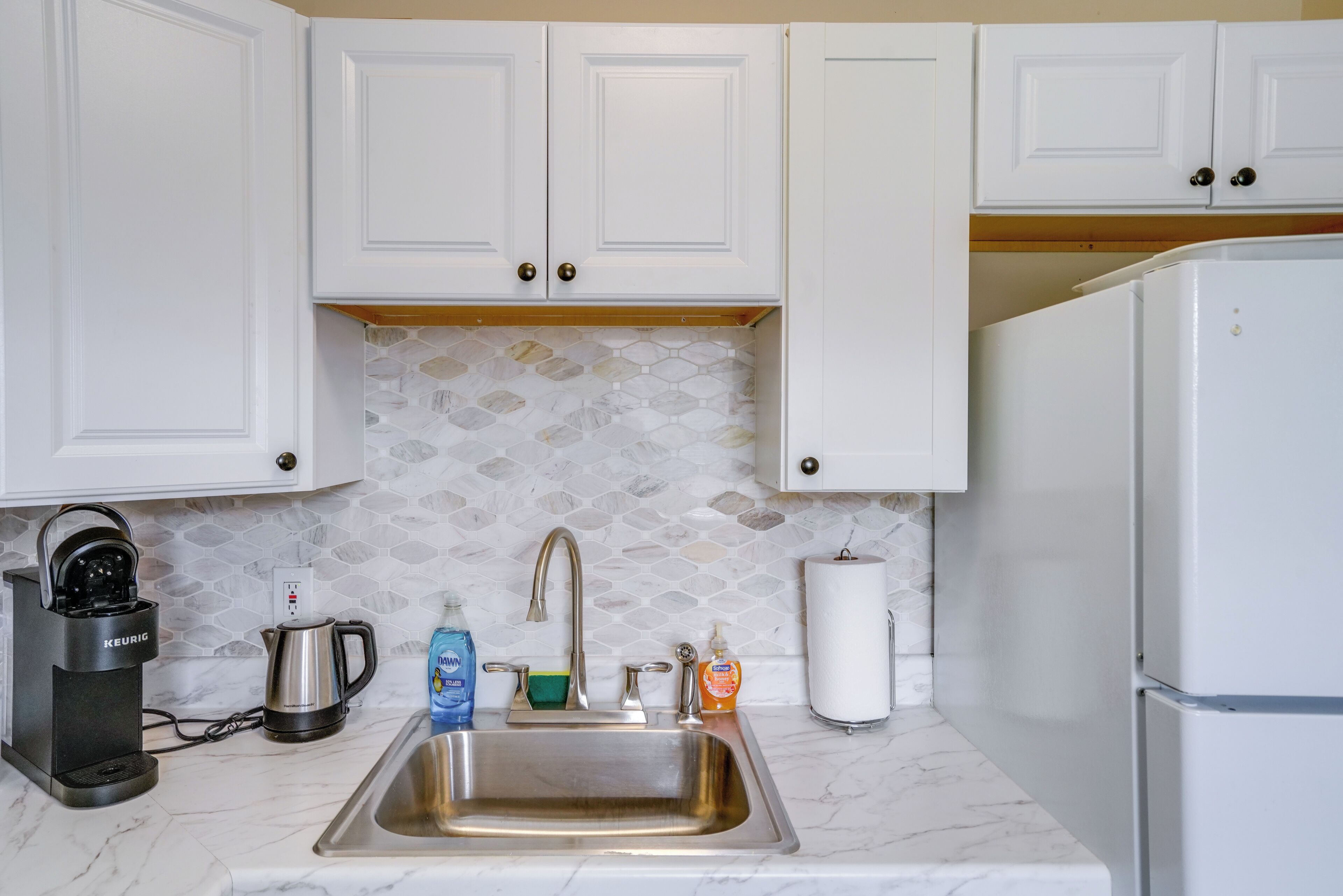 A kitchen with white cabinets, a stainless steel sink, a coffee maker, a kettle, a dish soap bottle, and a paper towel dispenser.