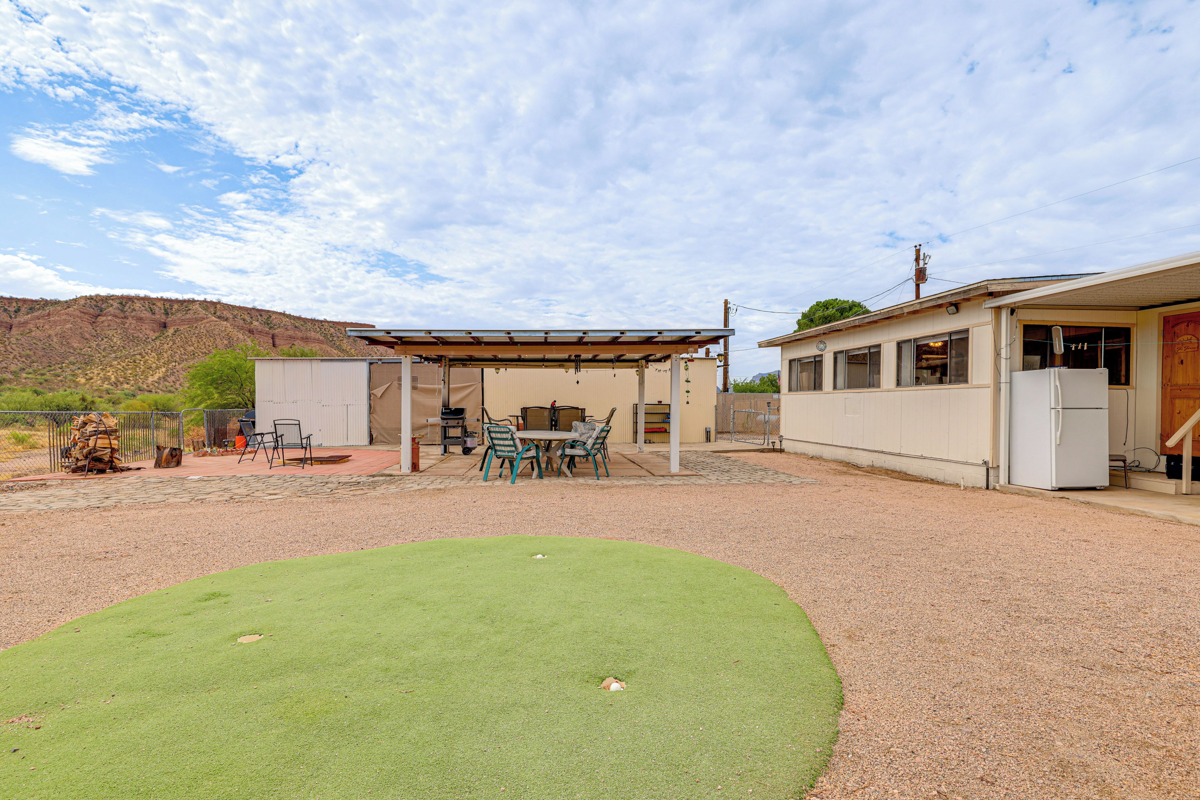 A covered outdoor area with a table and chairs, a barbecue grill, and a building with a refrigerator.