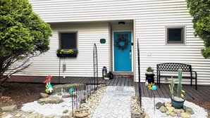 A white house with a blue door, a front porch with a bench, and a small garden with rocks and potted plants.