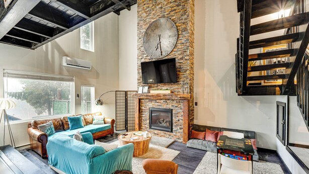 A modern living room with a stone fireplace, a large clock, and a staircase with exposed beams.
