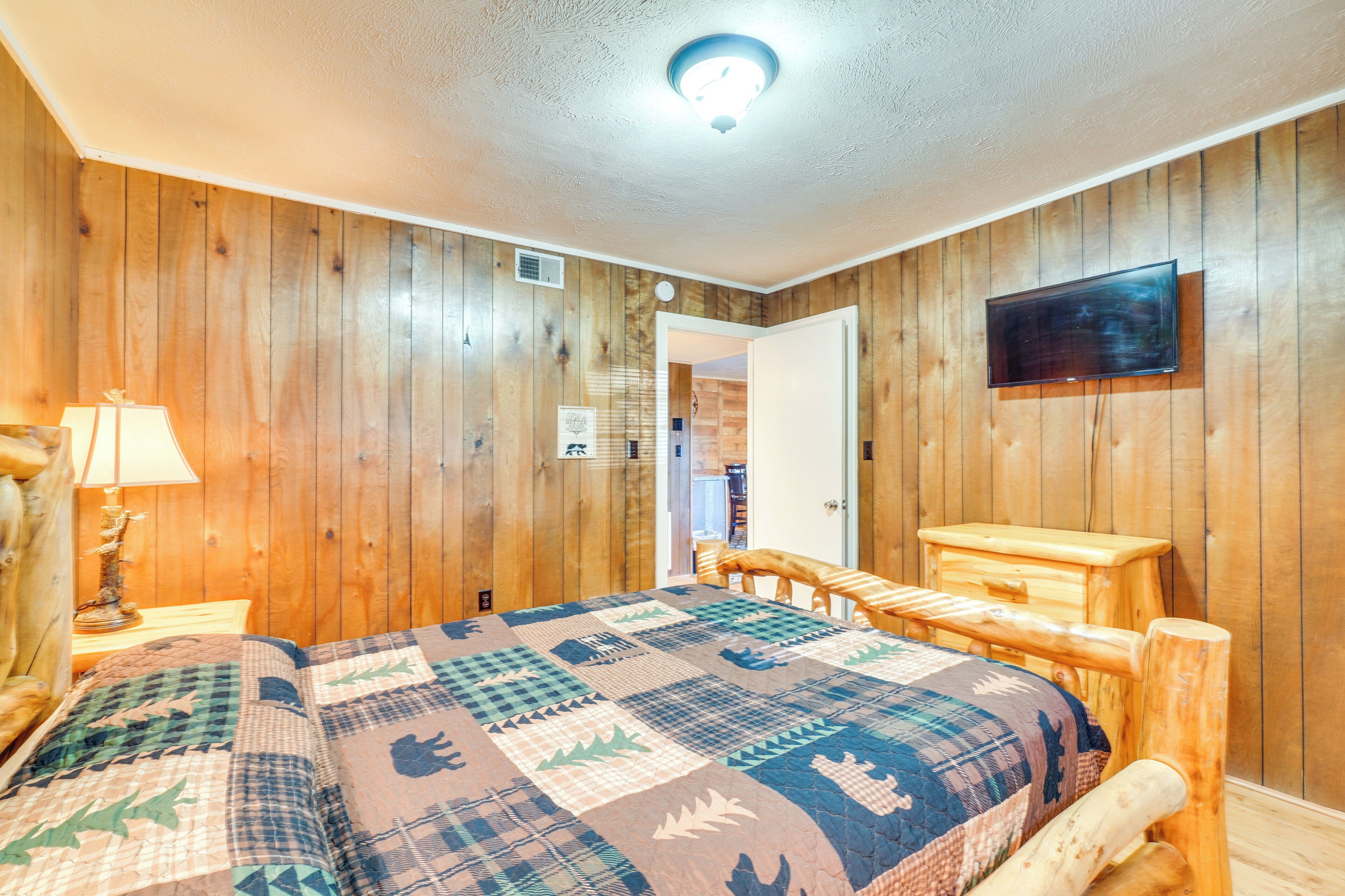 A wooden-paneled room with a bed, a nightstand, a lamp, a wooden chair, and a television mounted on the wall.