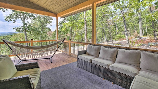 A covered outdoor seating area with a hammock, wicker sofa, and a view of a forested landscape. - 'the Glabin: Garrison Gem w/ Hot Tub & Fire Pit (Garrison)