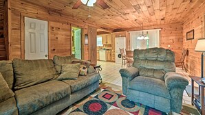 A wooden cabin interior with a sofa, armchair, ceiling fan, and a kitchen area visible through an open door. - Group-friendly Cabin on Kentucky Lake! (Springville)