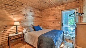 A wooden cabin bedroom with a bed, a nightstand, a lamp, and a window with blinds. - Group-friendly Cabin on Kentucky Lake! (Springville)