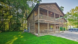 A two-story wooden cabin with a balcony on the upper floor, surrounded by a grassy yard and trees. - Group-friendly Cabin on Kentucky Lake! (Springville)