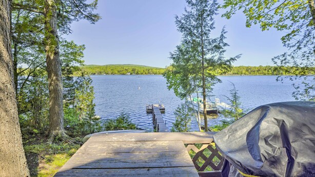 Une terrasse en bois donnant sur un lac, avec un quai et un bateau, le tout entouré d’arbres.