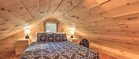 A bedroom with a sloped ceiling, wooden walls, a bed with a patterned bedspread, two bedside tables with lamps, and a window with blinds.
