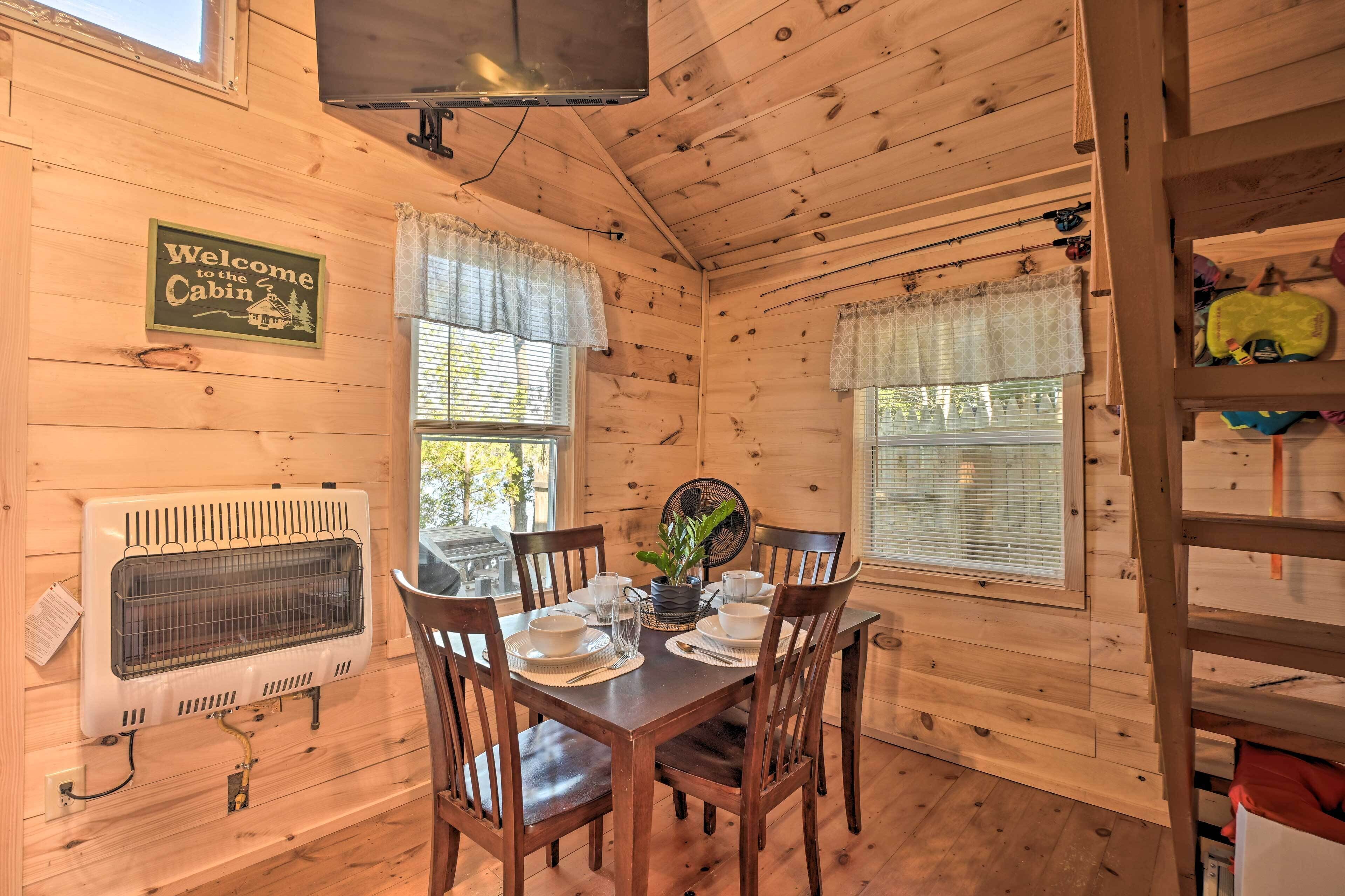 A wooden cabin interior with a dining table set for four, a television mounted on the ceiling, and a 'Welcome to the Cabin' sign.