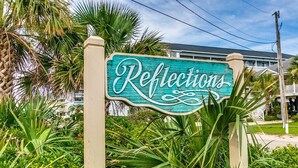 Um letreiro com a palavra 'Reflections' em fonte cursiva, cercado por vegetação exuberante.