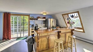 A kitchen with wooden cabinets, a stainless steel refrigerator, a wooden bar counter with stools, and a window with hanging pots. - Charming Studio ~ 20 Mi to Acadia Nat'l Park! (Ellsworth)