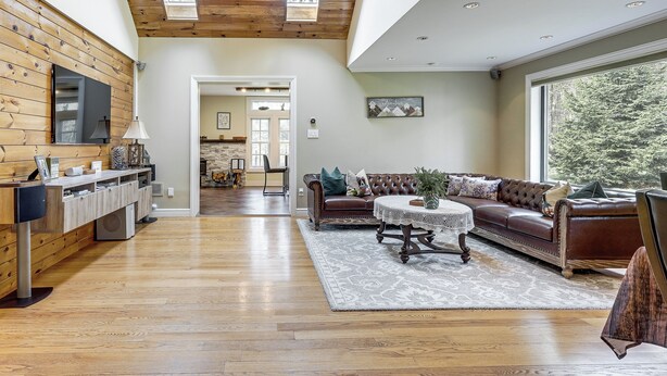 A spacious living room with a wooden ceiling, a large window, a brown leather sofa, and a dining area in the background. - Poconos Cottage w/ Sauna ~ 10 Mi to Camelback! (Tobyhanna)