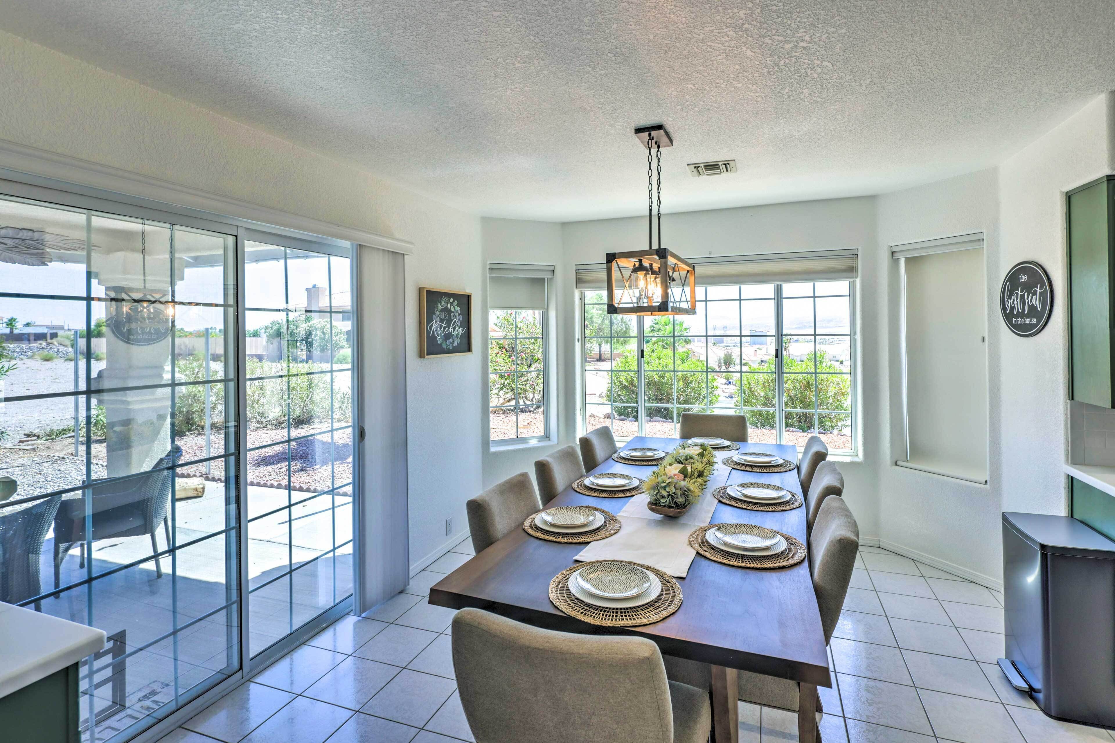 A dining area with a large table set for six, a hanging light fixture, and a view through large glass doors.