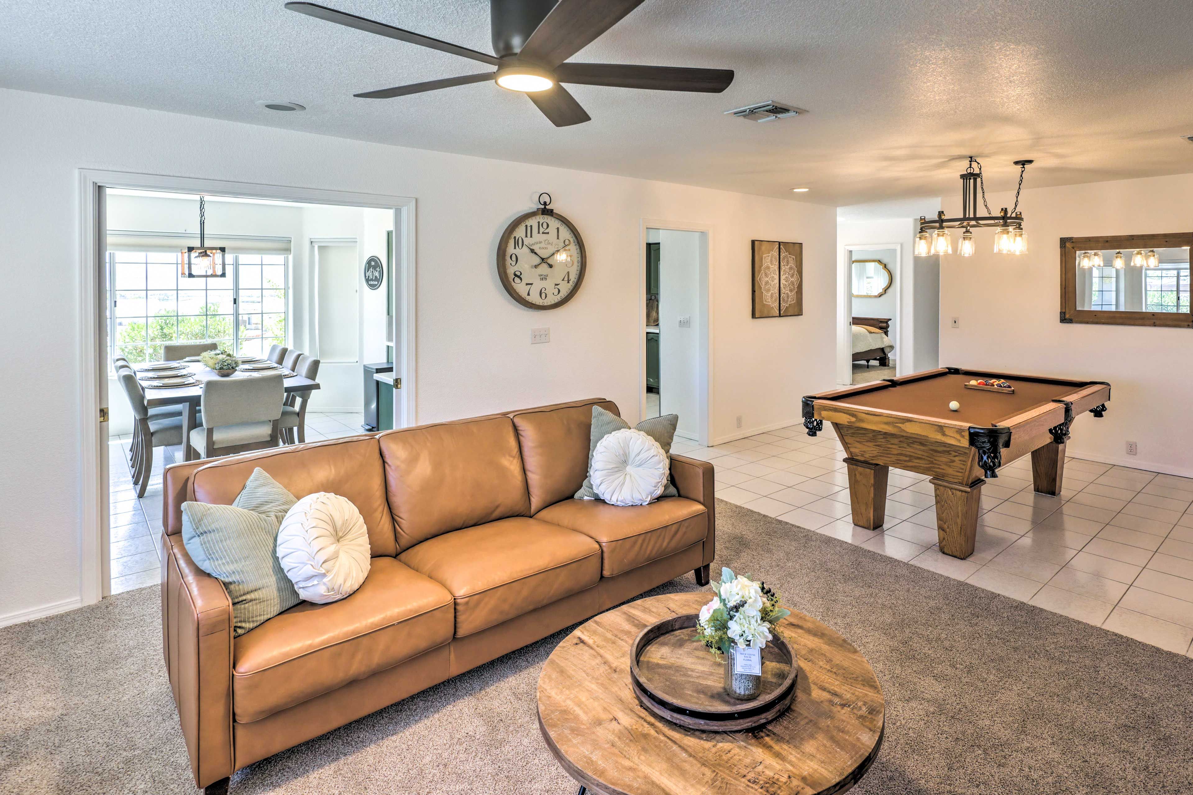 A living room with a brown leather sofa, a round coffee table with a vase, a wooden pool table, and a ceiling fan.