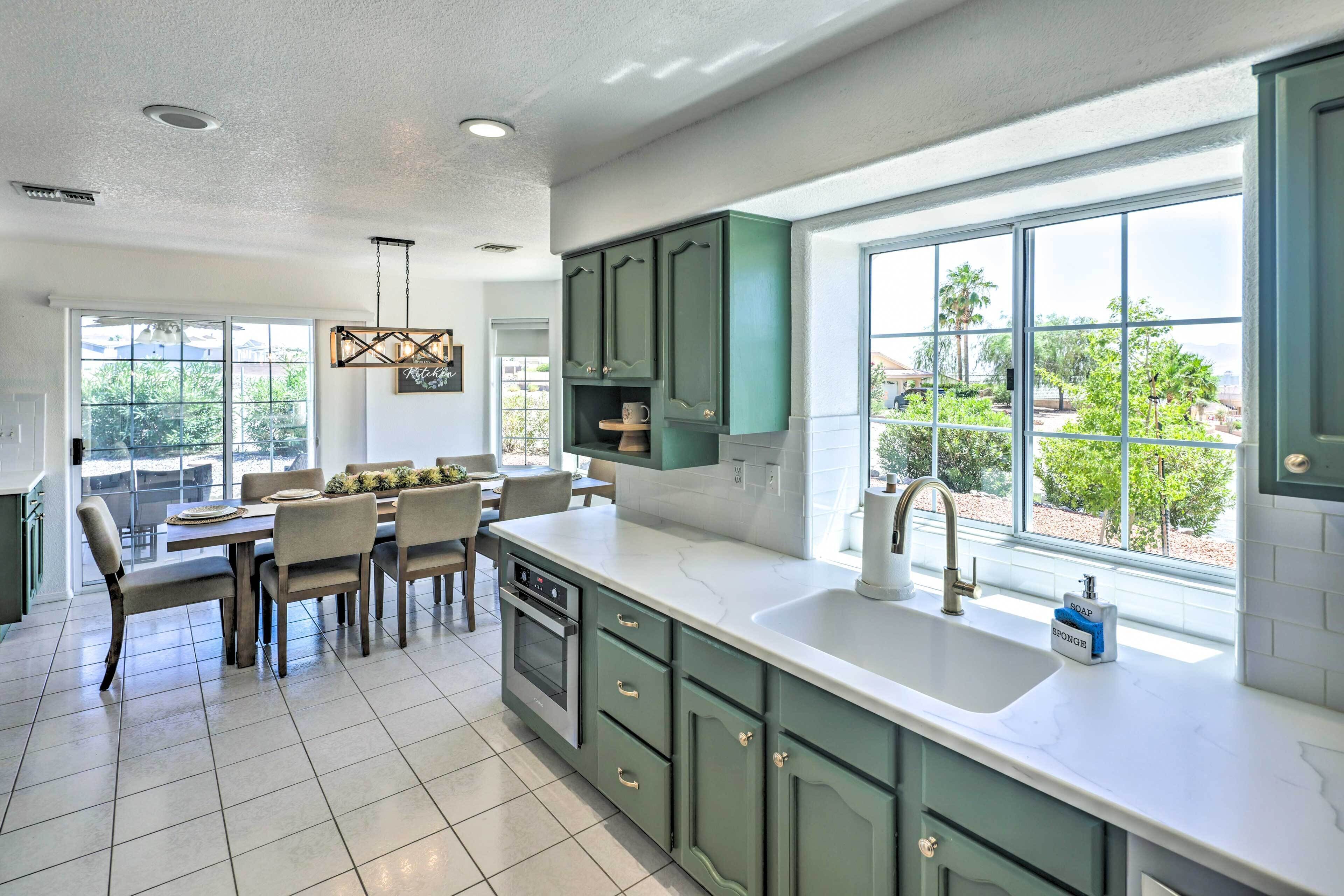 A modern kitchen with a large island, a dining area with chairs, and a view of greenery outside.