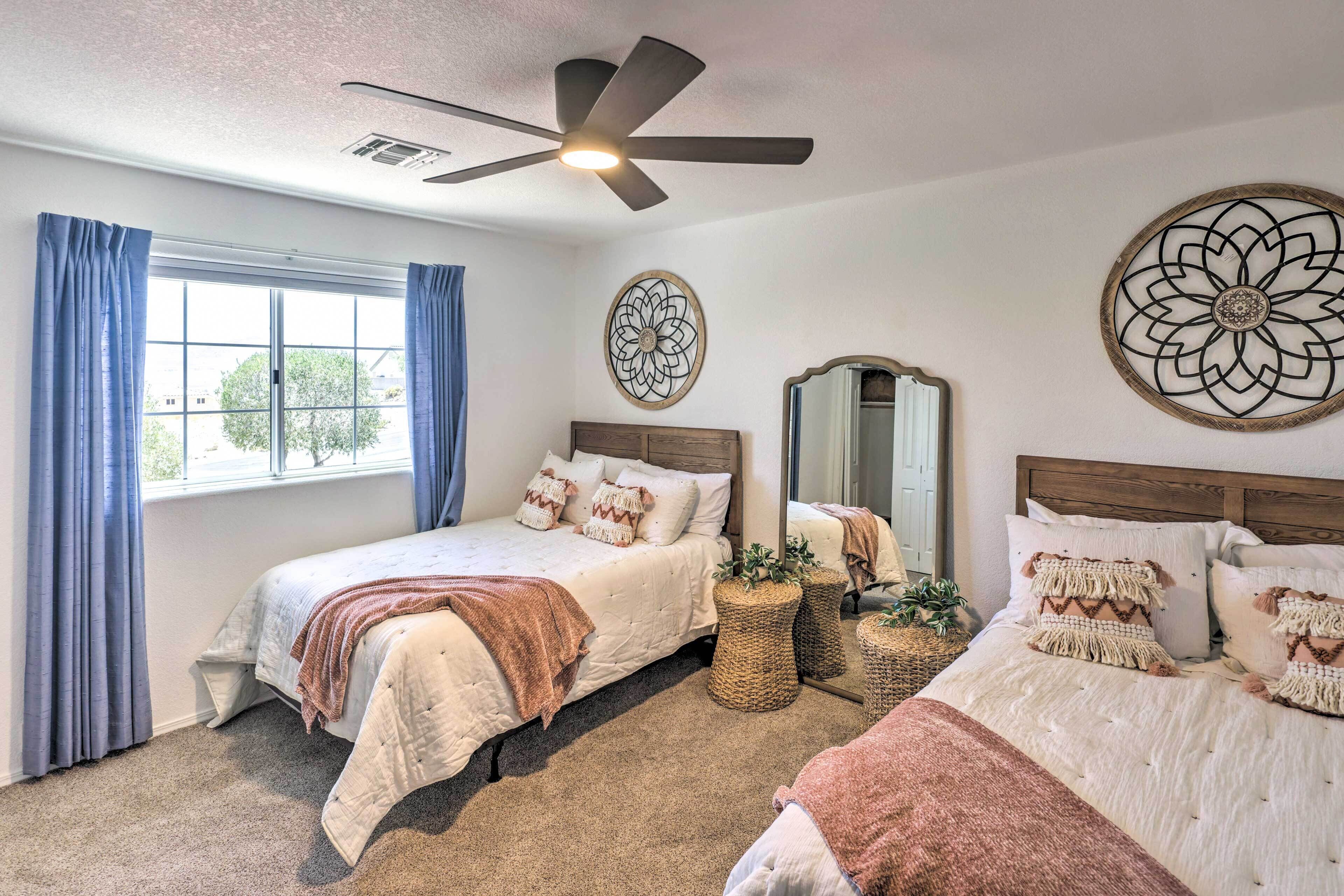 A bedroom with two beds, a ceiling fan, a large window with curtains, and decorative wall art.