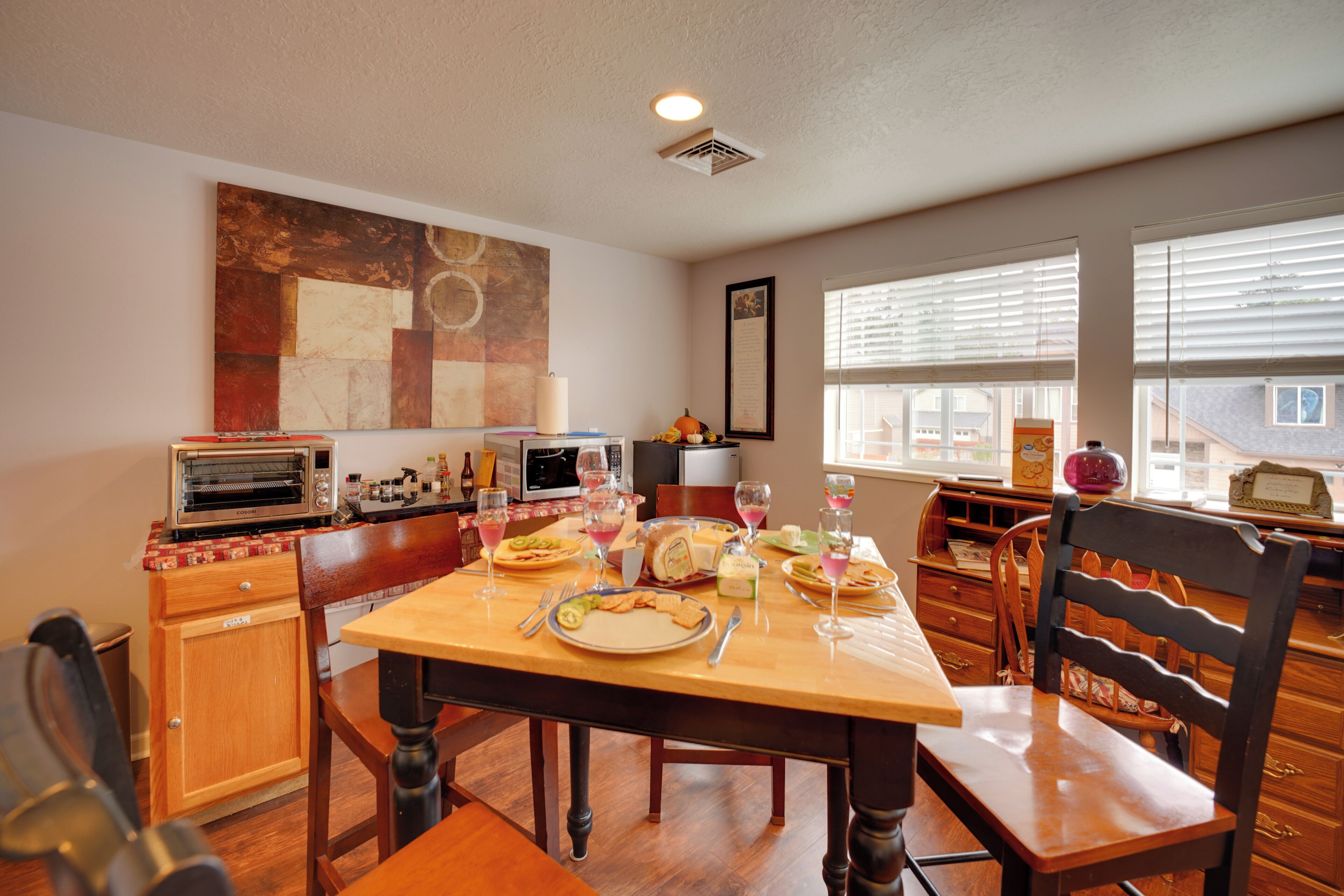 Uma sala de jantar com mesa de madeira posta para refeição, área de cozinha com eletrodomésticos e um grande quadro abstrato na parede.