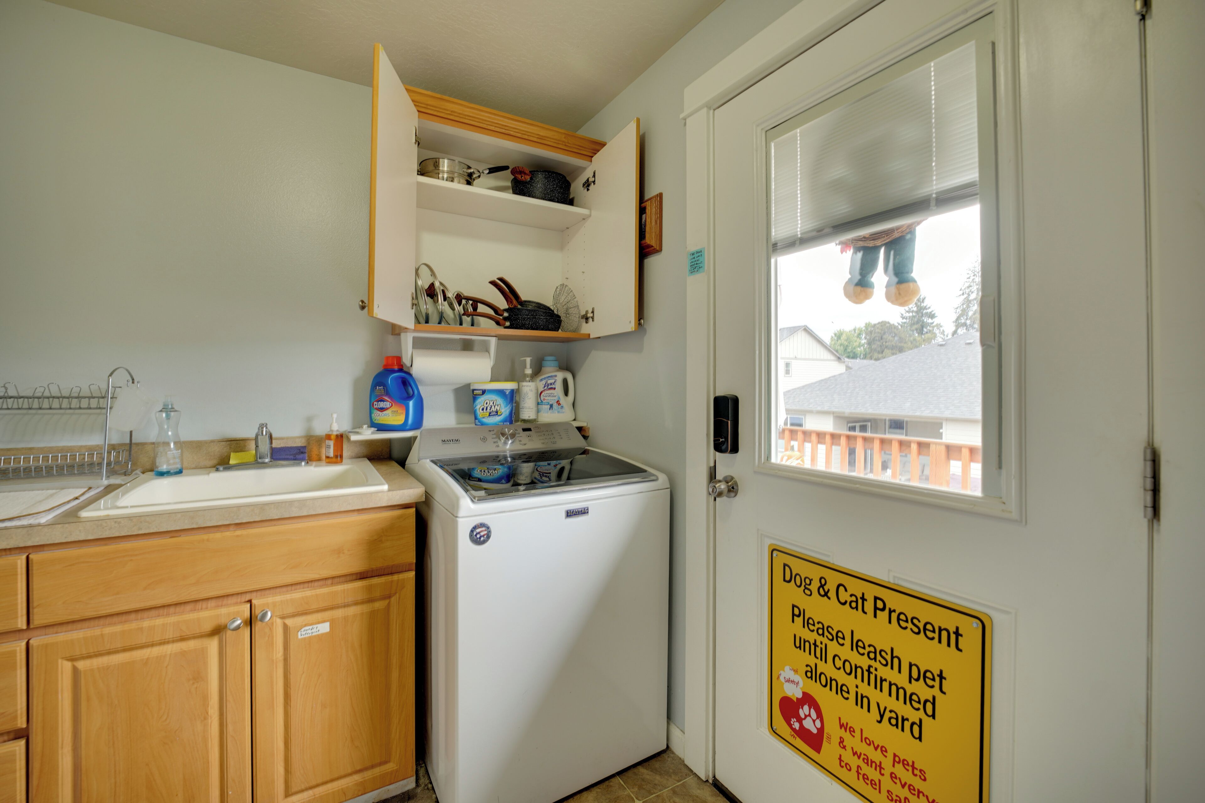 Une cuisine équipée d’un lave-linge, d’un évier et d’un placard contenant divers objets. Un panneau est affiché sur la porte demandant aux propriétaires de tenir leurs animaux en laisse.