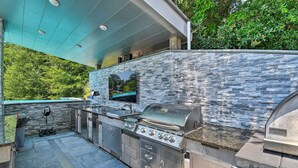 A modern outdoor kitchen with a stone backsplash, built-in grill, and a flat-screen TV. - Southern Shores Retreat w/ Expansive Outdoor Space (Kitty Hawk)