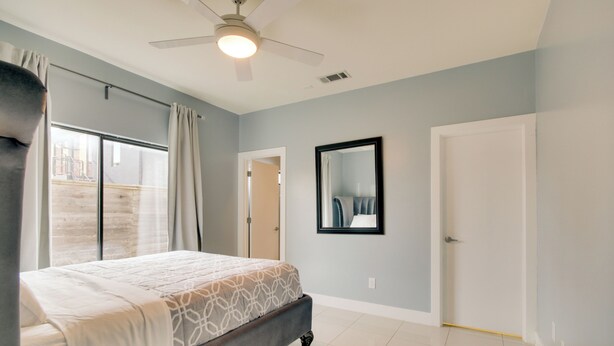 Une chambre à coucher avec un lit, un ventilateur de plafond, une grande fenêtre avec des rideaux et une porte.