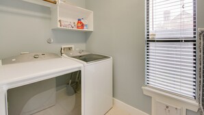 Un espace buanderie comprenant un lave-linge et un sèche-linge, une étagère avec des produits de lessive et une fenêtre avec des stores.