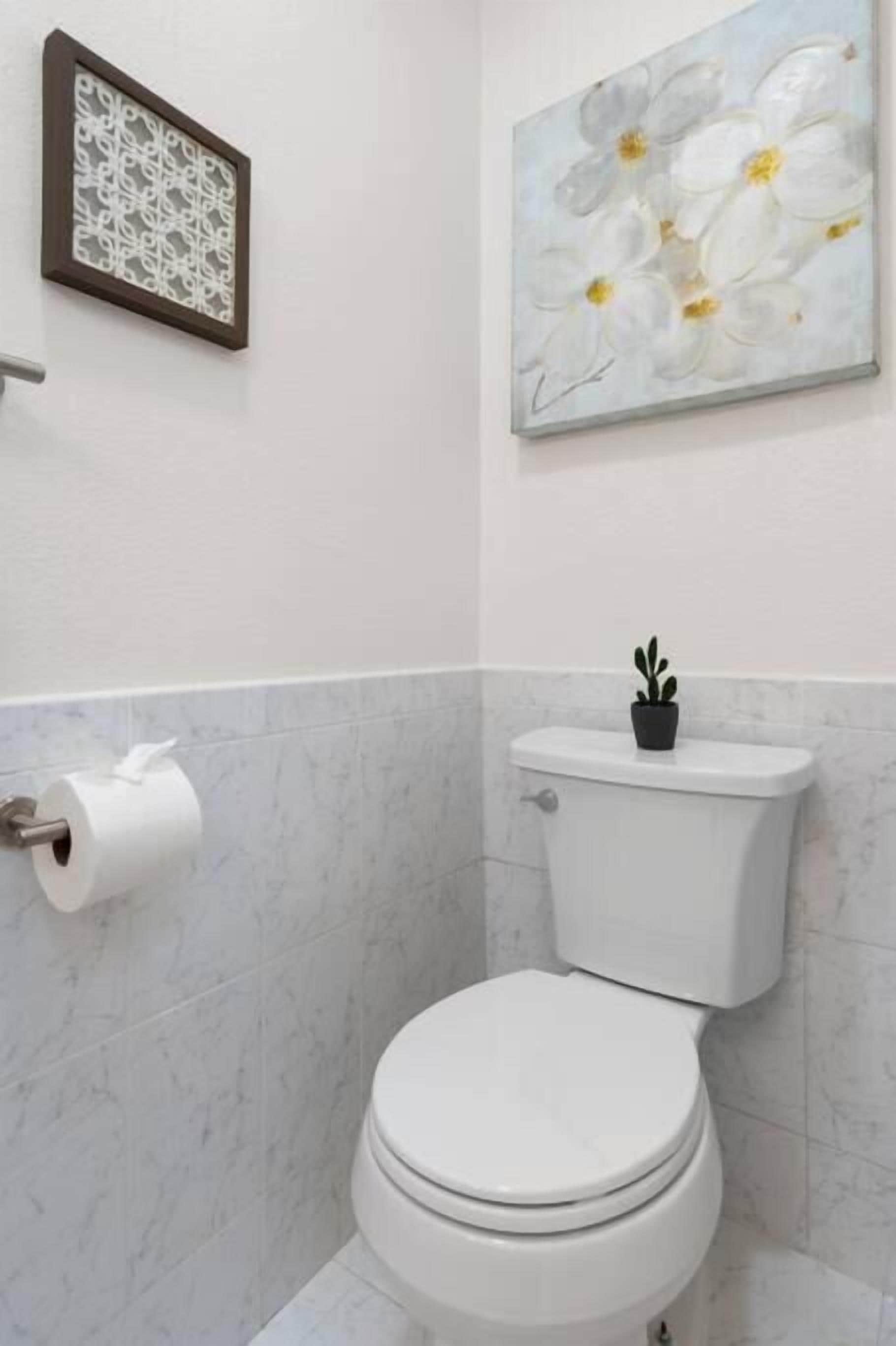 A modern bathroom with a toilet, a toilet paper holder, a framed abstract art piece, and a potted plant on the toilet tank.