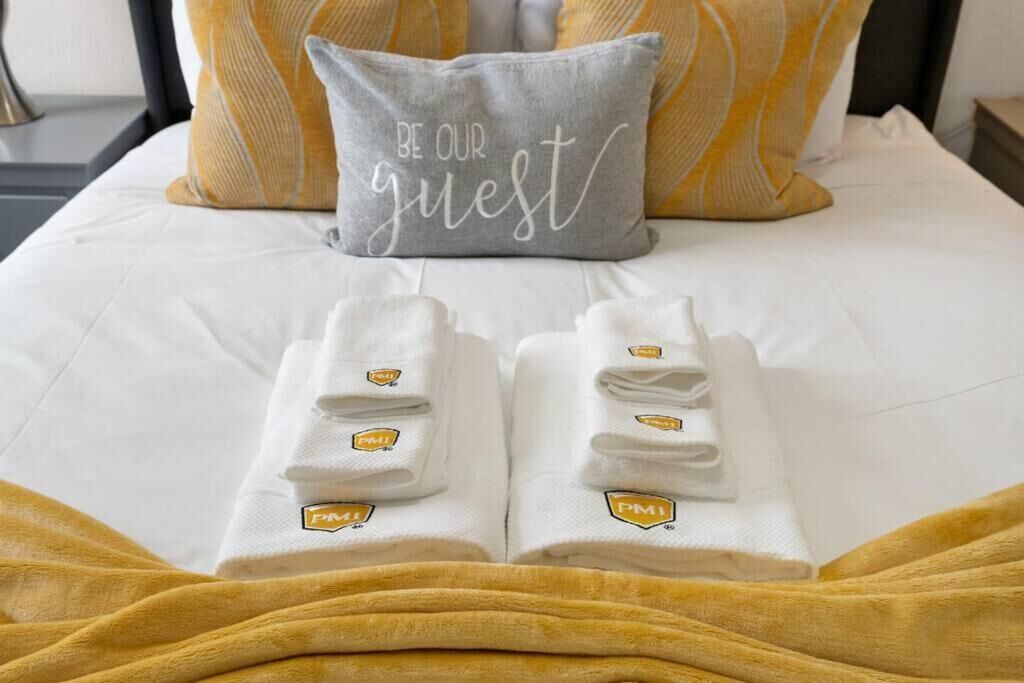 A neatly made bed with a pillow that reads "BE OUR guest" and two folded towels with a logo on them.
