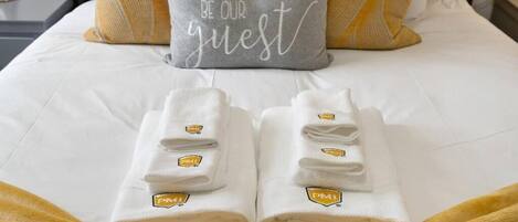 A neatly made bed with a pillow that reads "BE OUR guest" and two folded towels with a logo on them.