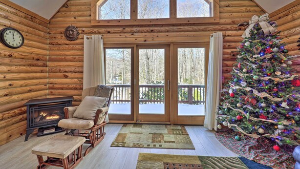 A log cabin interior with a fireplace, a Christmas tree, and a large window overlooking a snowy deck. - 2 Decks & Fire Pit: Scenic Beech Mountain Retreat! (Banner Elk)