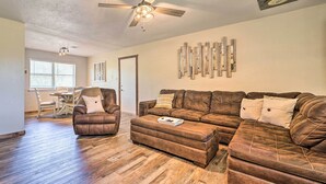 A living room with a brown leather sectional sofa, a matching ottoman, and a brown leather armchair. There is a ceiling fan, a dining table with chairs, and a window with blinds. - Private Hot Tub & Yard: Secluded Broken Bow Home! (Broken Bow)