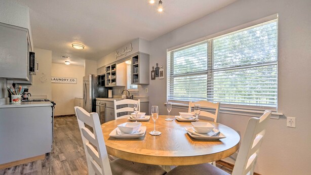 A dining area with a round wooden table set for four, surrounded by white chairs. A kitchen with white cabinets and a refrigerator is visible in the background. - Private Hot Tub & Yard: Secluded Broken Bow Home! (Broken Bow)