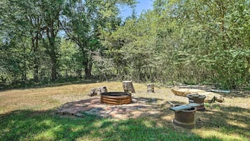 A rustic outdoor setting with a fire pit, stone benches, and a brick fire ring surrounded by trees.