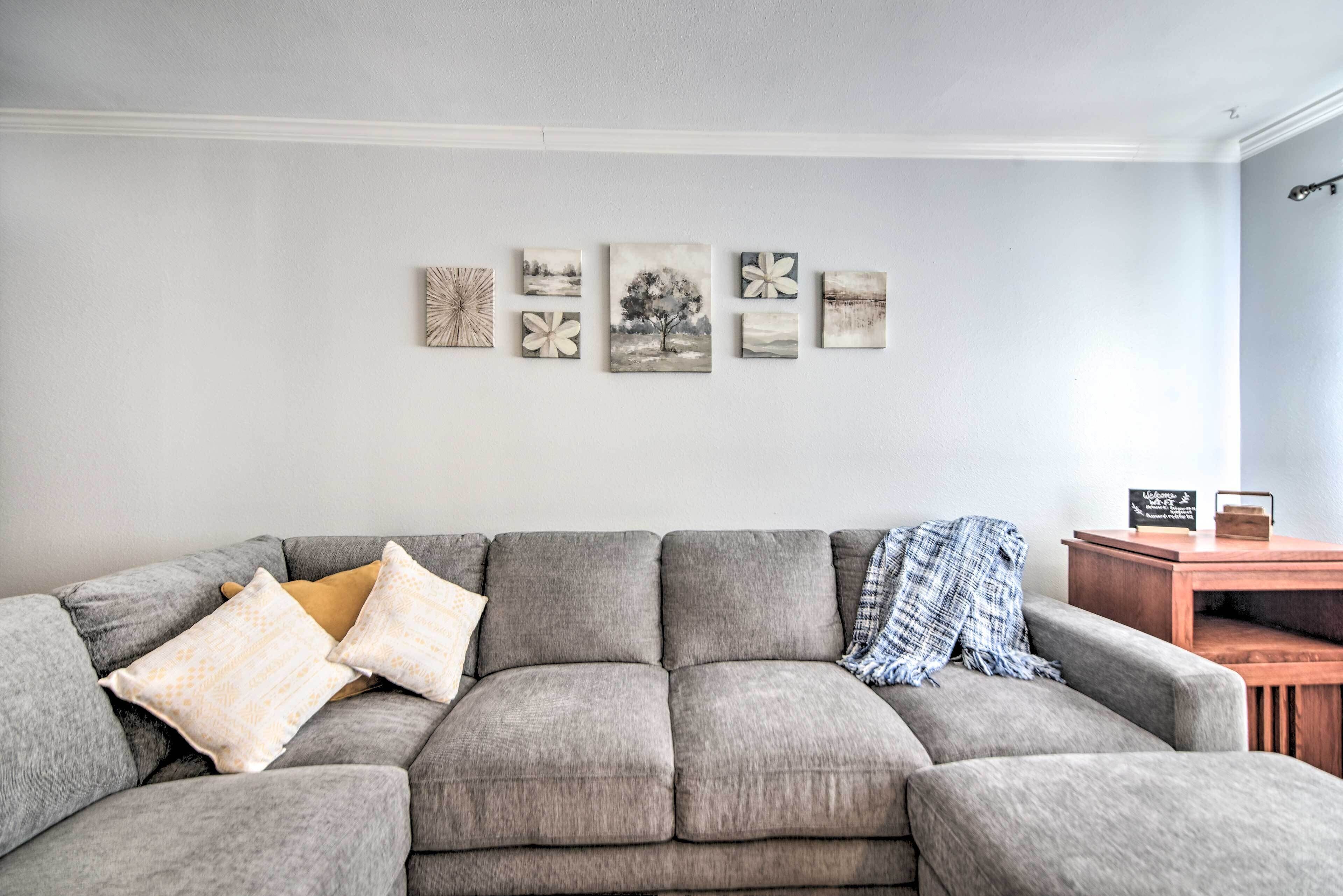 A grey sofa with patterned cushions, a wooden side table, and a wall with five framed artworks.