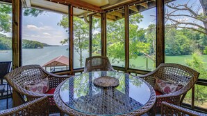 Une table en verre entourée de chaises en osier avec des coussins, de grandes fenêtres donnant sur un lac et des arbres.