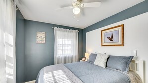 Ein Schlafzimmer mit Bett, Deckenventilator, Fenster mit Vorhängen, einem gerahmten Bild an der Wand und einem motivierenden Zitat.