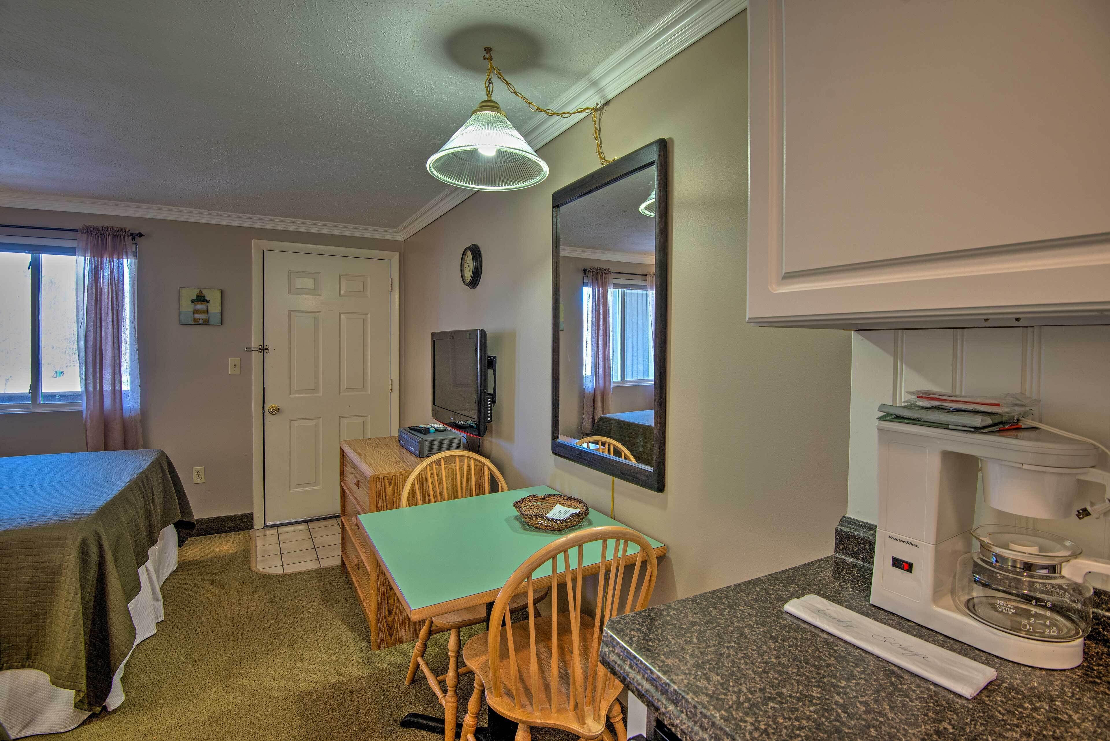 A compact room with a bed, a dining table with chairs, a kitchenette, a large mirror, and a ceiling light fixture.