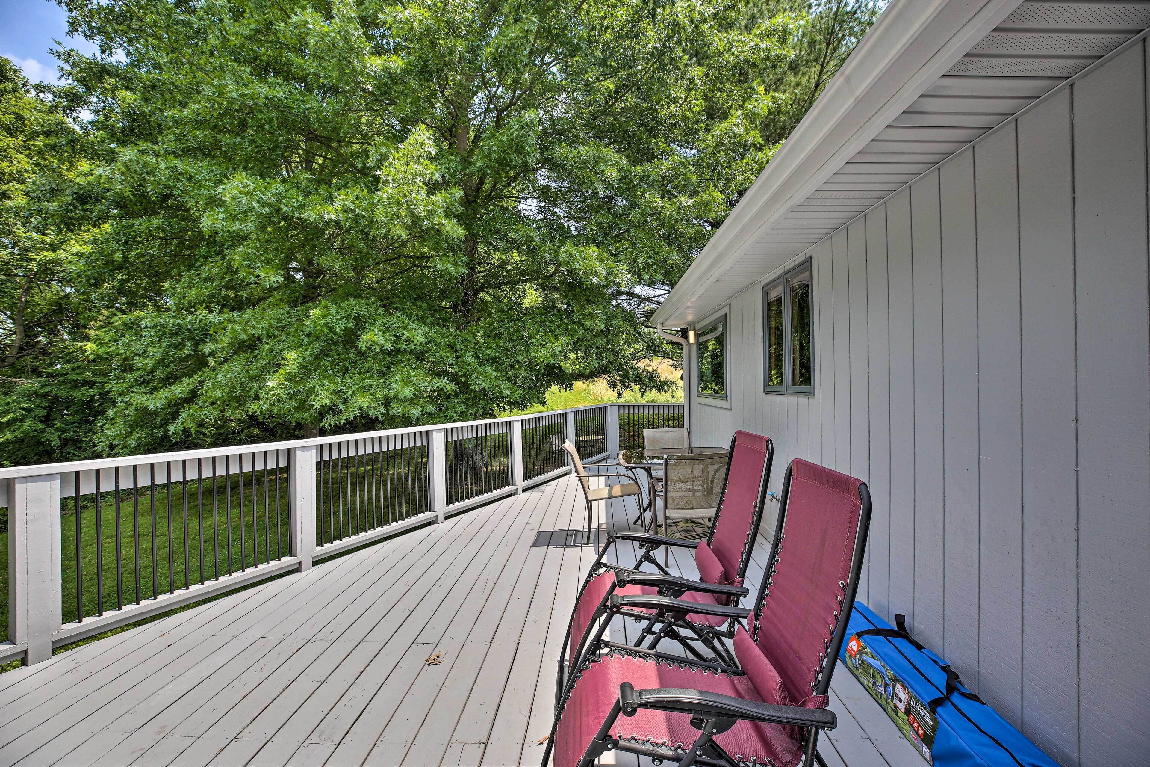 A wooden deck with a white railing, a covered seating area with chairs, and a view of a green landscape.