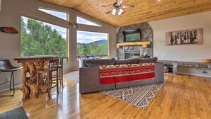 A spacious living room with a wooden ceiling, a stone fireplace, a large window with a view of trees, and a cozy seating area.