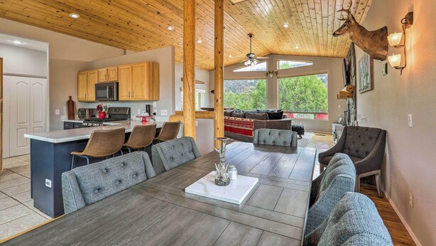A spacious living area with a large wooden ceiling, a dining table with grey upholstered chairs, and a mounted deer head on the wall.