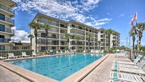 A swimming pool with lounge chairs and palm trees, surrounded by a multi-story building with balconies. - Ormond Beach Condo w/ Balcony & Views (Ormond Beach)