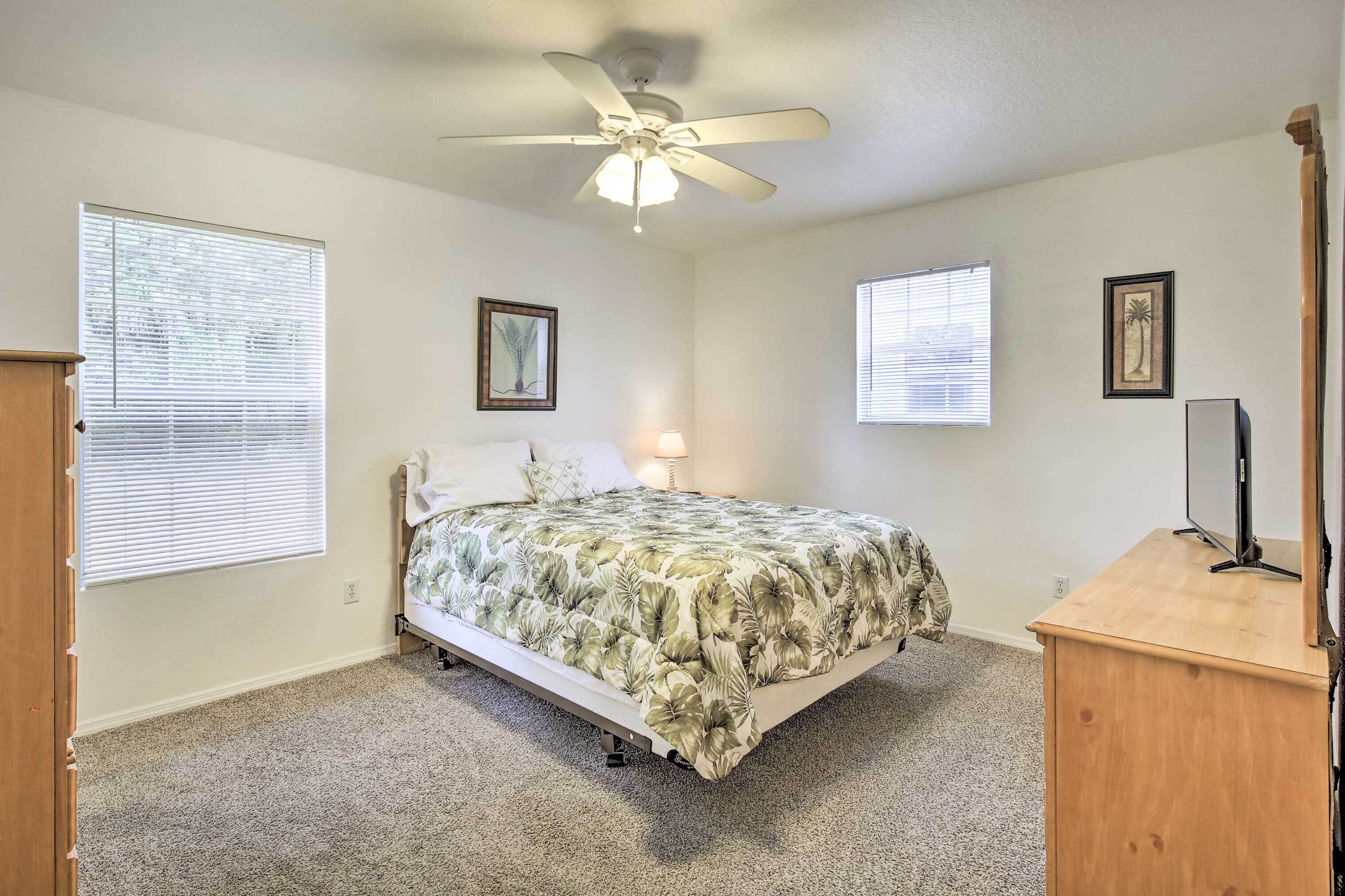 A bedroom with a bed, a ceiling fan, a desk, and two windows with blinds.