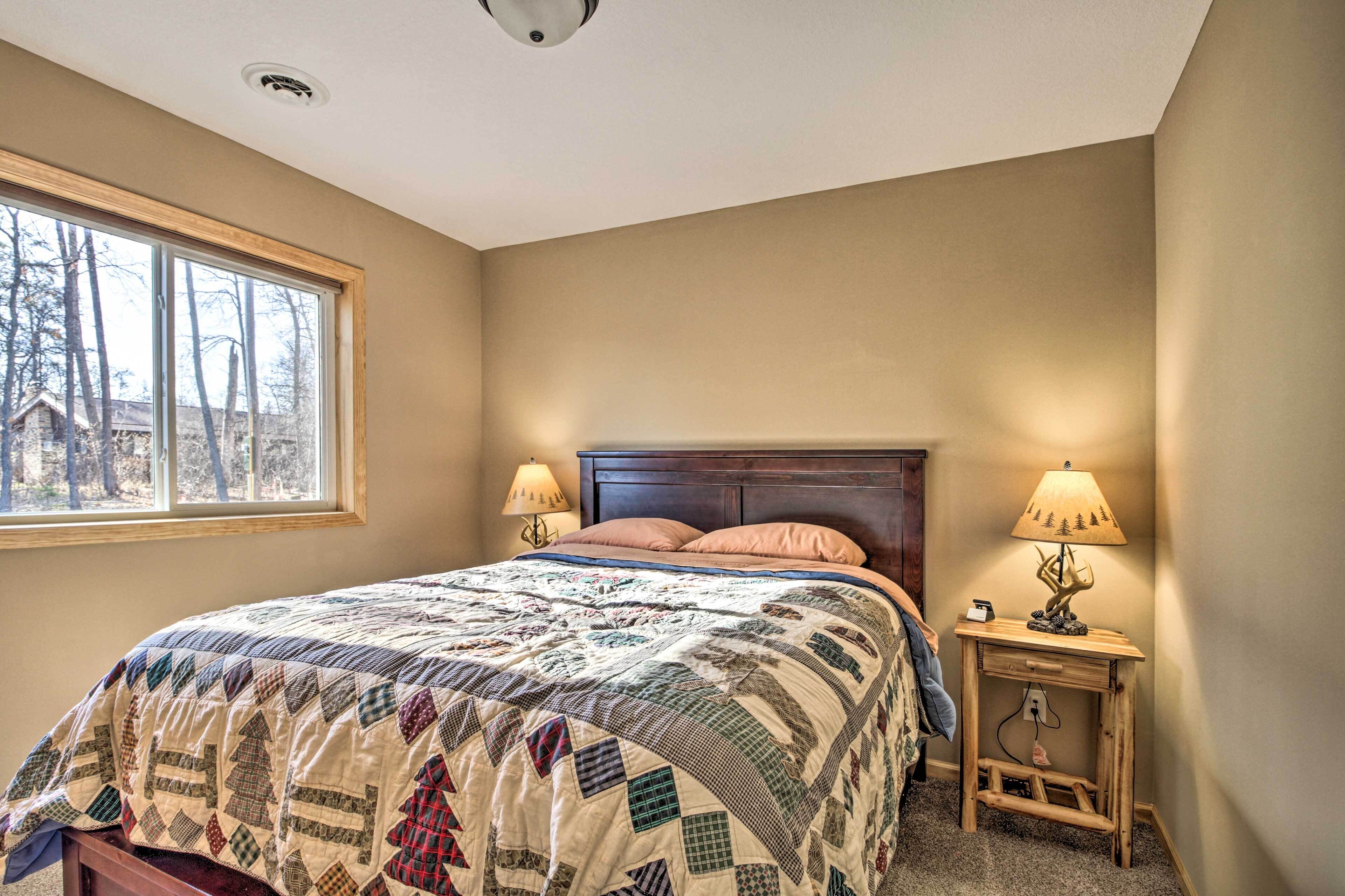 A bedroom with a large bed, two bedside lamps, a nightstand, and a window with a view of trees and a house.