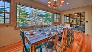 A dining area with a wooden table set for six, large windows with a forest view, and a bookshelf in the background. - Log Cabin in Wilmington w/ New Hot Tub By Skiing! (Wilmington)